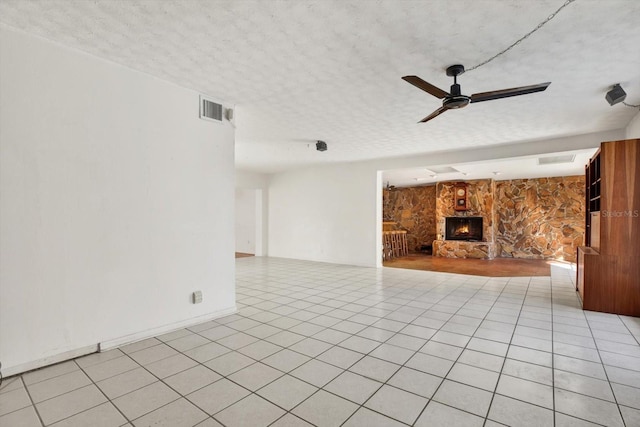 unfurnished living room with a textured ceiling, a stone fireplace, light tile patterned flooring, visible vents, and a ceiling fan