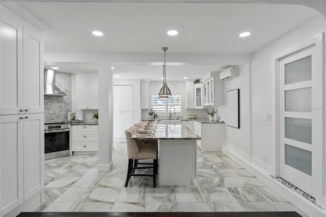 kitchen featuring a breakfast bar, white cabinets, an AC wall unit, wall chimney range hood, and stainless steel range with electric stovetop