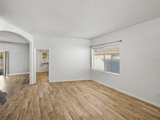 empty room with light wood-style flooring, a textured ceiling, arched walkways, and baseboards