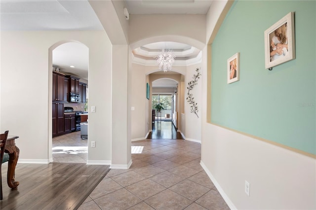 hall featuring arched walkways, crown molding, a raised ceiling, light tile patterned flooring, and baseboards