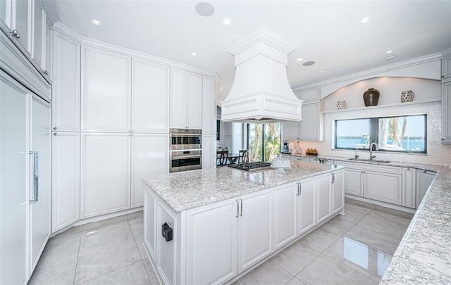 kitchen featuring light stone counters, premium range hood, a sink, white cabinetry, and a center island