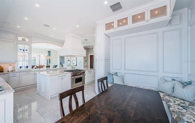 kitchen with visible vents, high end range, custom exhaust hood, white cabinetry, and a decorative wall