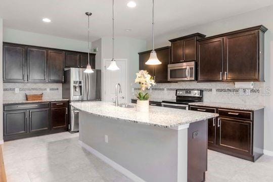 kitchen with light stone counters, an island with sink, a sink, dark brown cabinetry, and appliances with stainless steel finishes