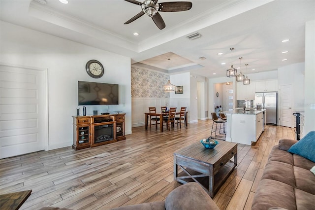 living room featuring a wainscoted wall, visible vents, light wood finished floors, a tray ceiling, and crown molding