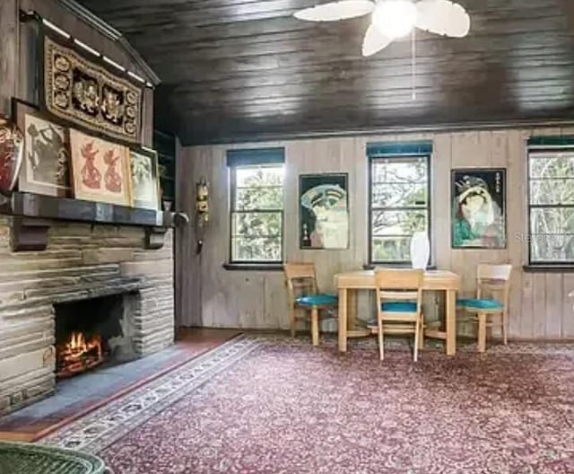 unfurnished room featuring a stone fireplace, wooden walls, a ceiling fan, wood ceiling, and carpet