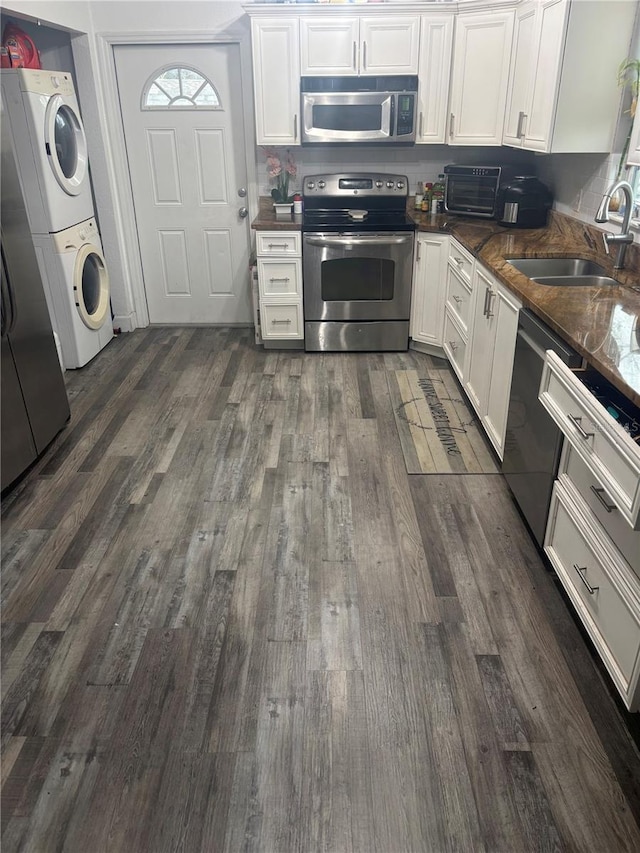 kitchen featuring stacked washer and clothes dryer, dark countertops, appliances with stainless steel finishes, white cabinetry, and a sink