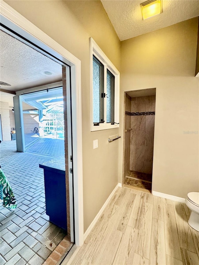 bathroom featuring baseboards, toilet, wood finished floors, a tile shower, and a textured ceiling