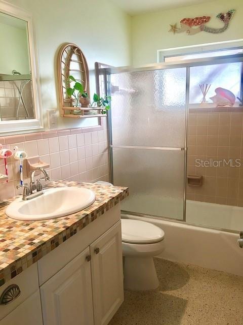 bathroom featuring toilet, speckled floor, vanity, tile walls, and combined bath / shower with glass door