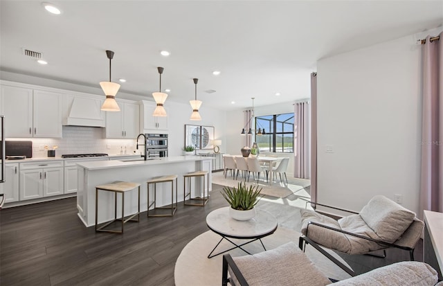 kitchen with a kitchen island with sink, gas stovetop, light countertops, backsplash, and custom exhaust hood