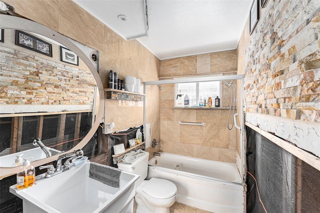 bathroom featuring toilet, shower / bath combination with glass door, brick wall, a textured ceiling, and a sink