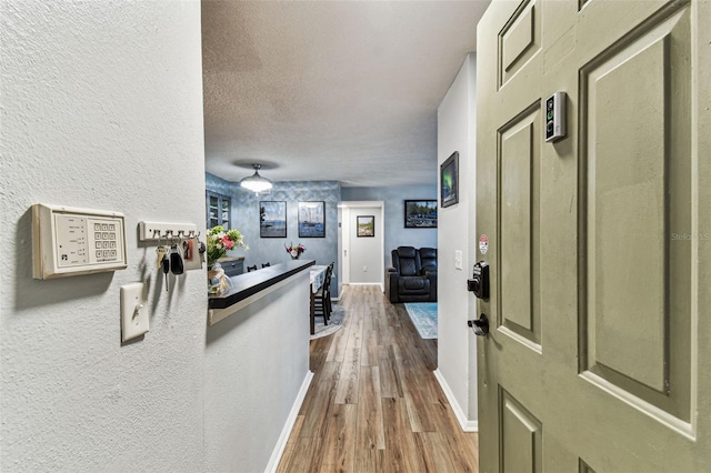 hall featuring a textured ceiling, a textured wall, wood finished floors, and baseboards