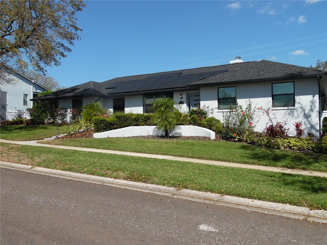 ranch-style house featuring solar panels, a front lawn, roof with shingles, and brick siding