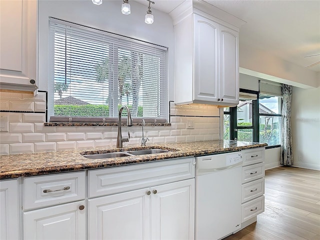 kitchen with white dishwasher, a sink, white cabinets, decorative backsplash, and dark stone counters