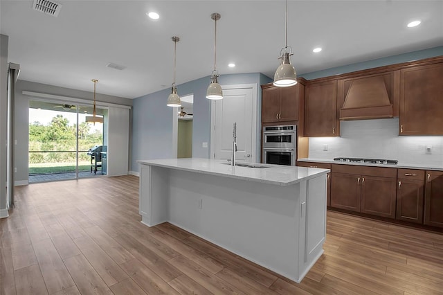 kitchen with stainless steel appliances, premium range hood, a sink, light wood-style floors, and decorative backsplash