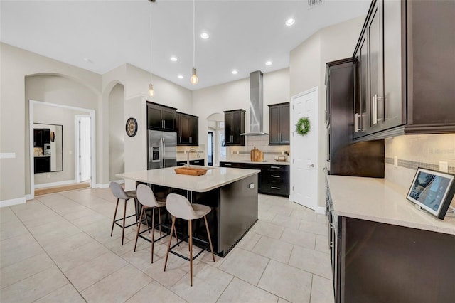 kitchen featuring a breakfast bar area, a kitchen island with sink, light countertops, wall chimney range hood, and stainless steel fridge