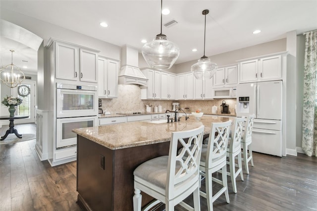 kitchen with white appliances, a center island with sink, visible vents, premium range hood, and backsplash