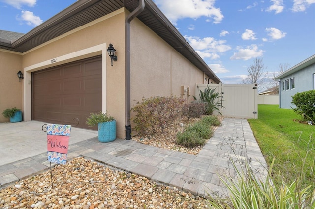 view of side of home featuring stucco siding, concrete driveway, an attached garage, a gate, and fence