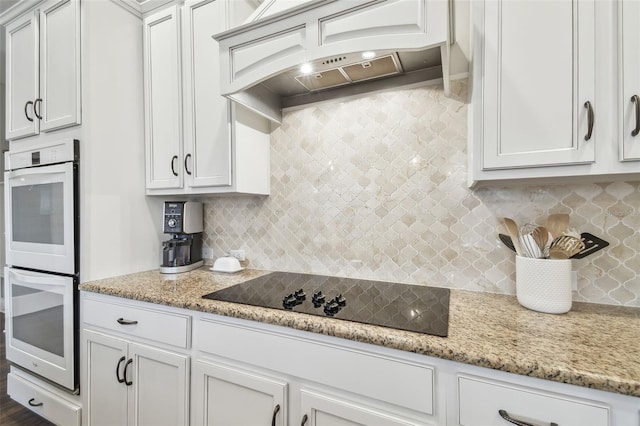 kitchen featuring black electric stovetop, white double oven, light stone counters, custom exhaust hood, and decorative backsplash