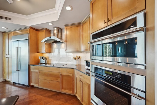 kitchen featuring dark wood finished floors, a tray ceiling, wall chimney range hood, visible vents, and stainless steel appliances
