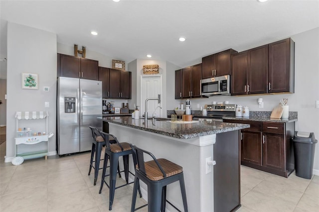 kitchen featuring a breakfast bar, appliances with stainless steel finishes, a sink, dark stone countertops, and dark brown cabinets