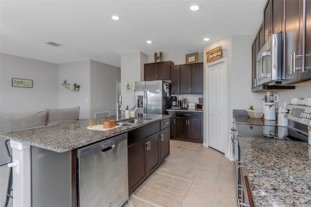 kitchen with stone counters, appliances with stainless steel finishes, a kitchen island with sink, a sink, and dark brown cabinets