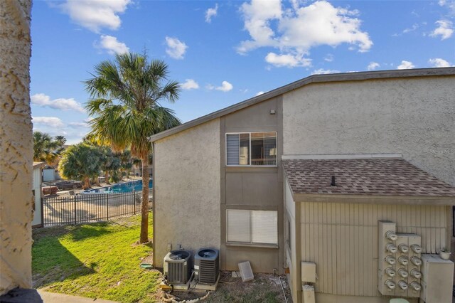 view of side of home with stucco siding, fence, central AC unit, and roof with shingles