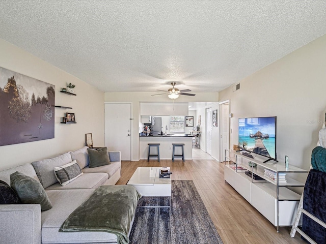 living area featuring light wood-type flooring, ceiling fan, visible vents, and a textured ceiling