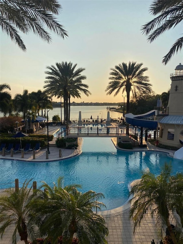 pool at dusk featuring a water slide, a water view, fence, and a community pool