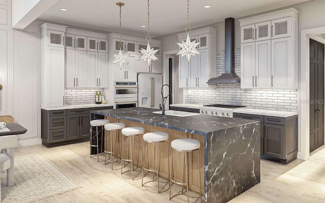 kitchen with white double oven, an island with sink, wall chimney exhaust hood, high end fridge, and a sink