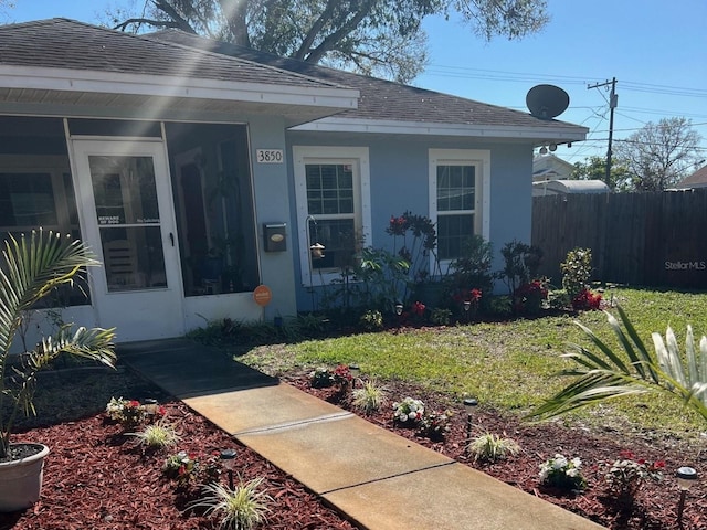 view of exterior entry featuring stucco siding, a lawn, roof with shingles, and fence