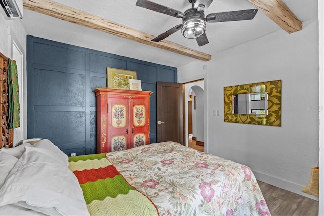 bedroom featuring arched walkways, beam ceiling, baseboards, and wood finished floors