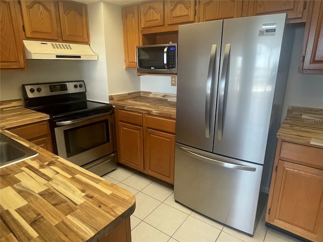 kitchen with light tile patterned floors, brown cabinetry, wood counters, appliances with stainless steel finishes, and under cabinet range hood