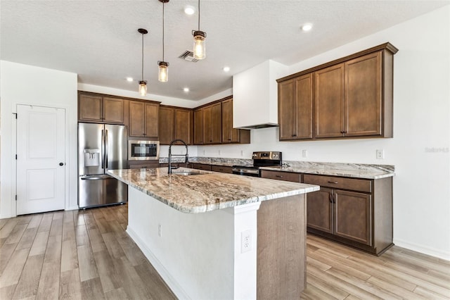 kitchen with appliances with stainless steel finishes, light wood-style floors, a sink, an island with sink, and light stone countertops