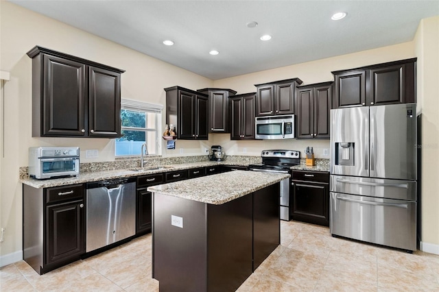 kitchen featuring a kitchen island, light stone counters, dark cabinets, stainless steel appliances, and a sink