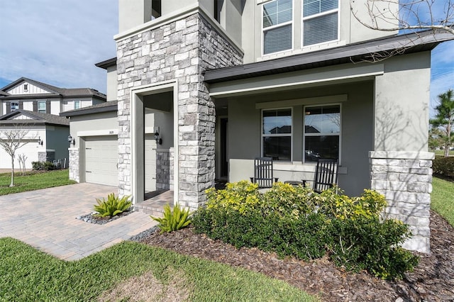 doorway to property featuring covered porch, a garage, stone siding, decorative driveway, and stucco siding