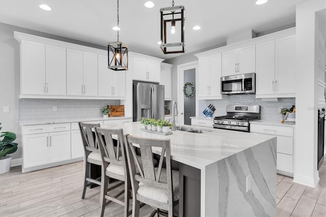 kitchen with a kitchen island with sink, stainless steel appliances, a sink, white cabinets, and decorative light fixtures