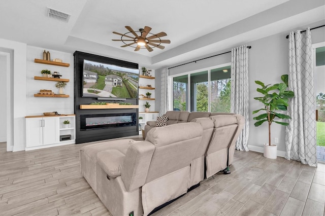 living area with light wood-style flooring, visible vents, a ceiling fan, a tray ceiling, and a glass covered fireplace