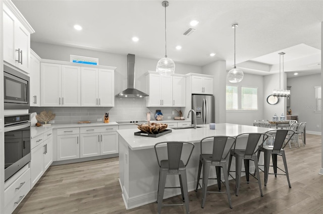 kitchen with visible vents, an island with sink, stainless steel appliances, wall chimney range hood, and backsplash