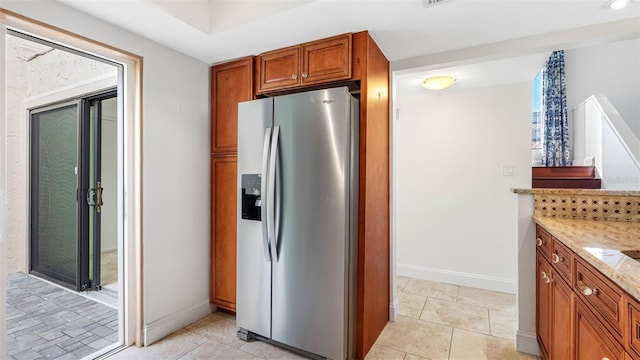 kitchen featuring light tile patterned floors, stainless steel fridge, baseboards, brown cabinets, and light stone countertops