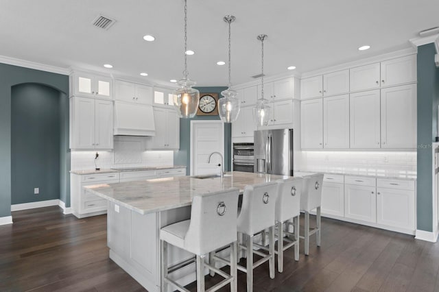 kitchen featuring arched walkways, a kitchen island with sink, stainless steel appliances, premium range hood, and white cabinetry