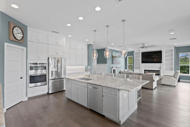 kitchen featuring a kitchen island with sink, stainless steel appliances, a sink, white cabinets, and a glass covered fireplace