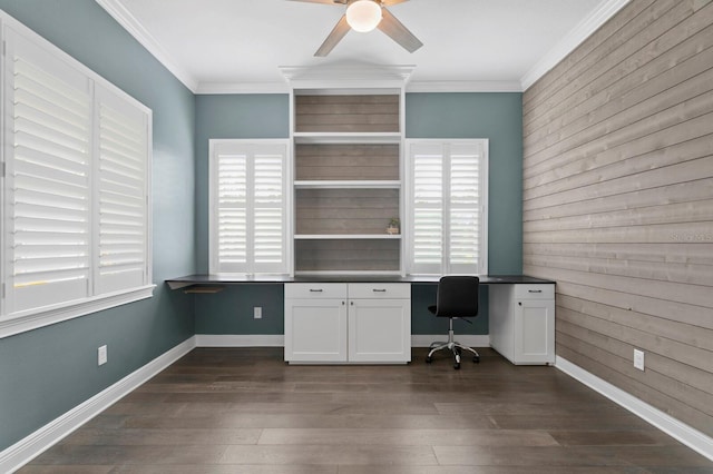 unfurnished office featuring crown molding, dark wood finished floors, built in desk, a ceiling fan, and baseboards