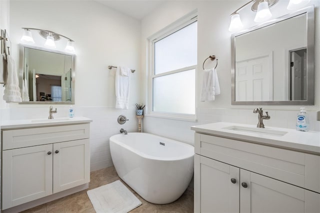 full bath featuring tile walls, a freestanding tub, two vanities, and a sink