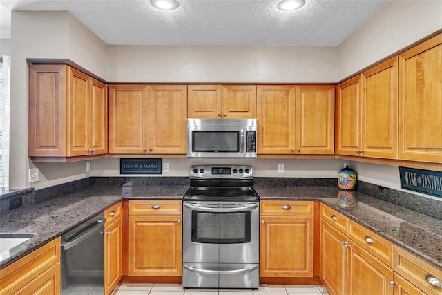 kitchen with stainless steel appliances, dark stone counters, brown cabinetry, and light tile patterned floors