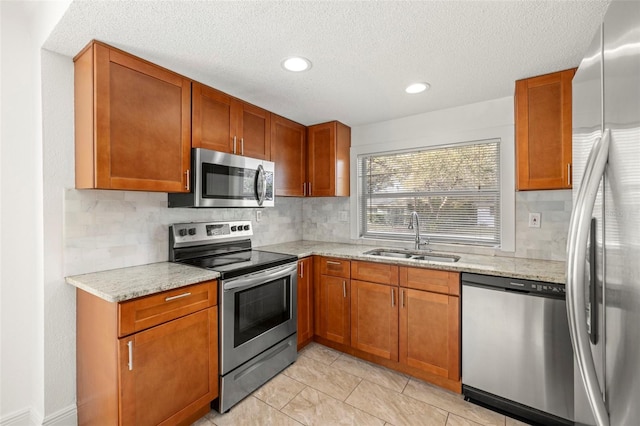 kitchen with stainless steel appliances, decorative backsplash, brown cabinetry, a sink, and light stone countertops