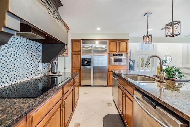 kitchen with brown cabinets, appliances with stainless steel finishes, a sink, dark stone counters, and under cabinet range hood