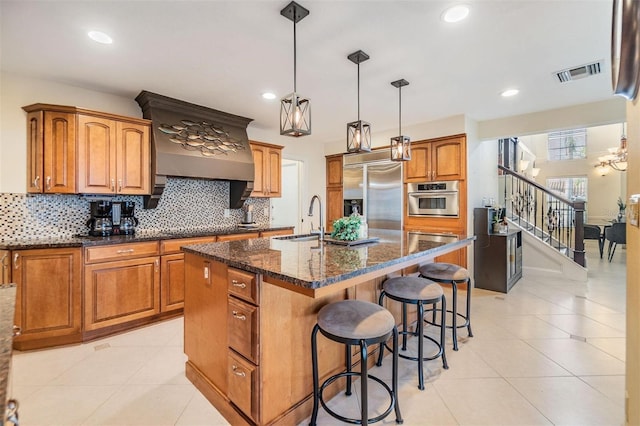 kitchen featuring stainless steel appliances, visible vents, brown cabinetry, dark stone countertops, and a center island with sink