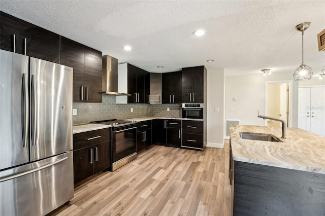 kitchen with light wood-style flooring, a sink, stainless steel appliances, wall chimney range hood, and decorative backsplash