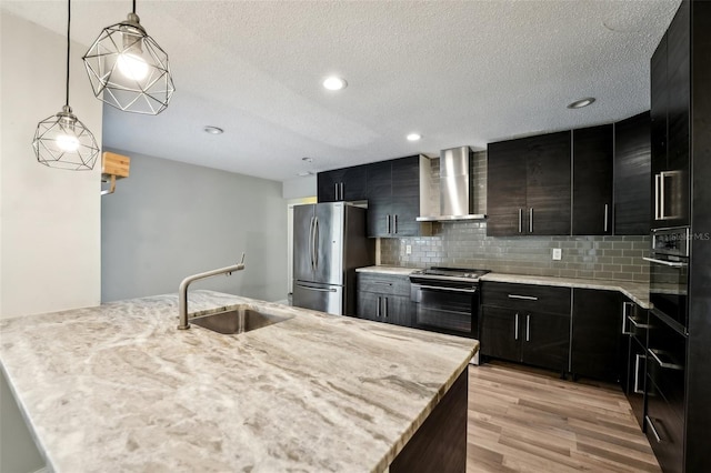 kitchen featuring light wood finished floors, appliances with stainless steel finishes, dark cabinetry, wall chimney exhaust hood, and a sink