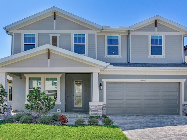 view of front of house with decorative driveway, roof with shingles, covered porch, an attached garage, and stone siding